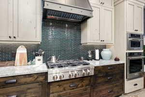 Kitchen with exhaust hood, backsplash, light stone counters, appliances with stainless steel finishes, and dark brown cabinets