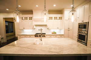 Kitchen featuring glass insert cabinets, a center island with sink, pendant lighting, light stone countertops, and recessed lighting