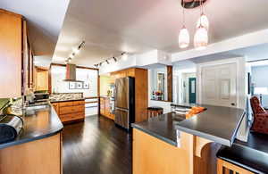 Kitchen with hanging light fixtures, brown cabinetry, dark countertops, stainless steel appliances, and a breakfast bar area