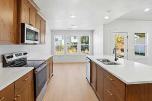 Kitchen with stainless steel appliances, brown cabinetry, light wood-style floors, recessed lighting, and a kitchen island with sink
