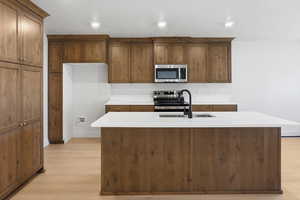 Kitchen with light wood-style flooring, appliances with stainless steel finishes, a center island with sink, brown cabinetry, and recessed lighting