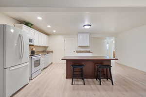 Kitchen with white appliances, white cabinetry, light wood-style flooring, recessed lighting, and a breakfast bar area