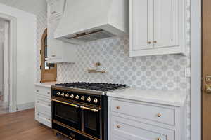 Kitchen with double oven range, custom exhaust hood, white cabinets, light stone countertops, and dark wood-style floors