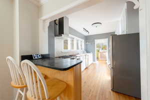 Kitchen with stainless steel appliances, dark countertops, white cabinets, light wood finished floors, and wall chimney exhaust hood