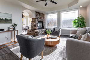 Living room with wood finished floors, a raised ceiling, arched walkways, a stone fireplace, and a ceiling fan