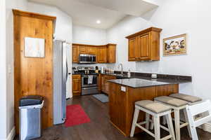 Kitchen featuring brown cabinetry, dark stone counters, stainless steel appliances, dark wood-style flooring, and a peninsula