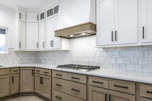 Kitchen featuring custom exhaust hood, white cabinets, glass insert cabinets, and tasteful backsplash