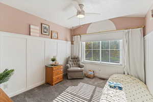 Bedroom featuring a decorative wall, dark carpet, a ceiling fan, wainscoting, and vaulted ceiling