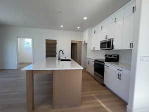 Kitchen with stainless steel appliances, white cabinets, light wood-type flooring, a center island with sink, and recessed lighting
