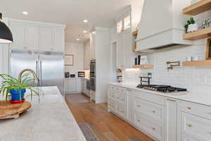 Kitchen with open shelves, appliances with stainless steel finishes, custom range hood, light stone countertops, and light wood finished floors
