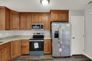 Kitchen with brown cabinetry, appliances with stainless steel finishes, light stone countertops, and dark wood finished floors