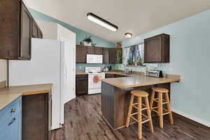 Kitchen with dark brown cabinetry, lofted ceiling, white appliances, light countertops, and a kitchen breakfast bar