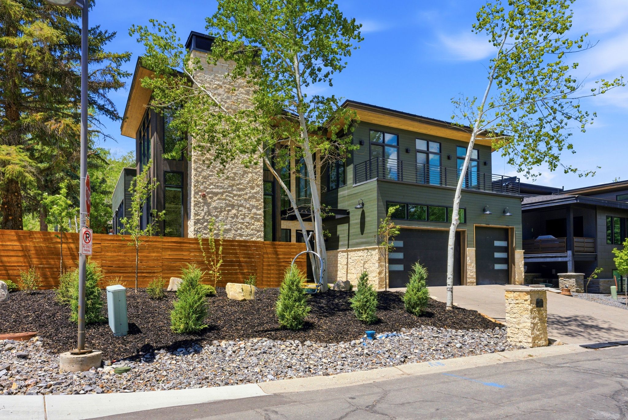 Modern exterior featuring stone and wood siding, a wooden privacy fence, a two-car garage with contemporary doors, and landscaped front yard with mulch and small evergreens