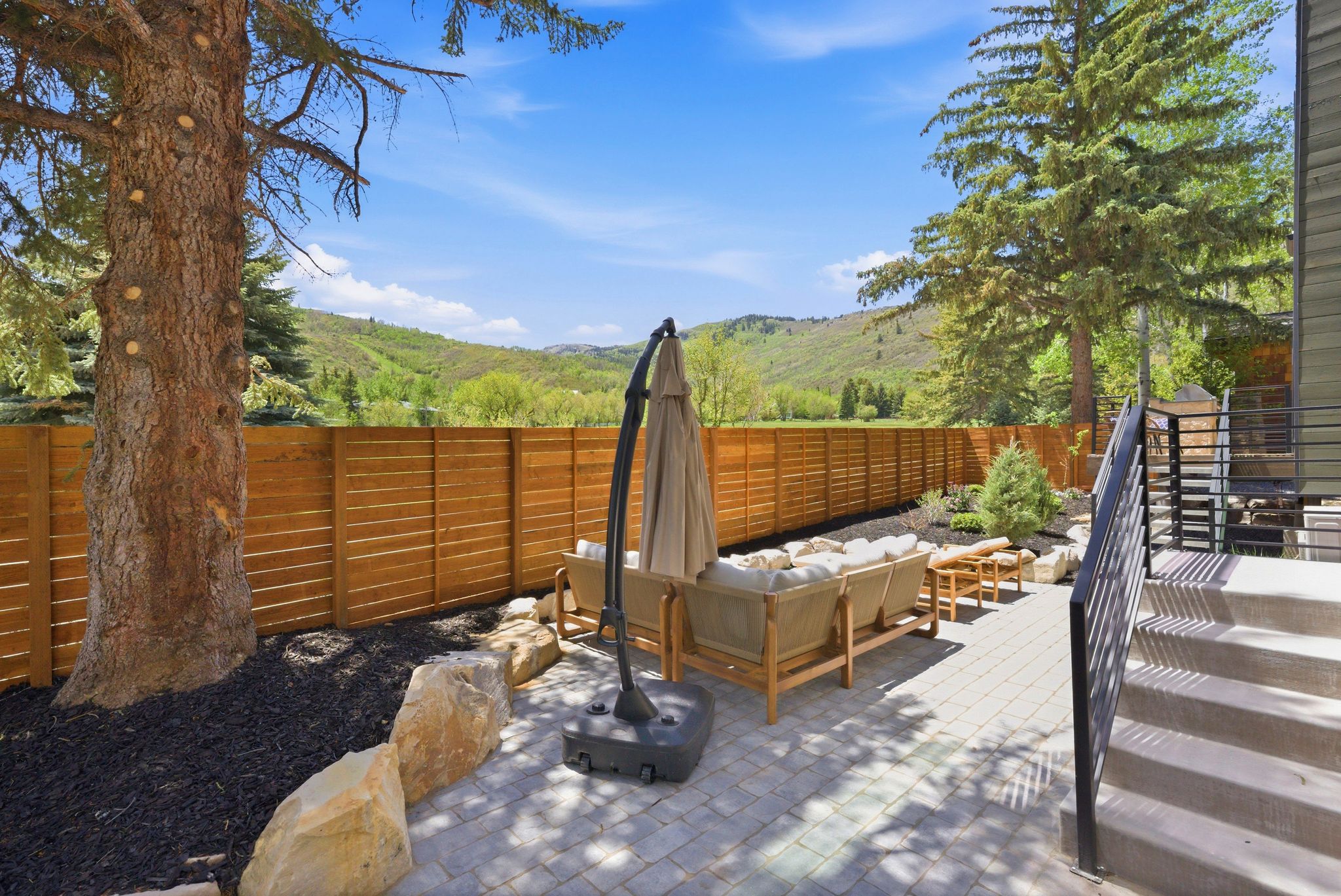 Paved patio area featuring paver flooring, a horizontal slat wood fence, mature landscaping with a large tree and decorative boulders, and exterior stairs with dark railings