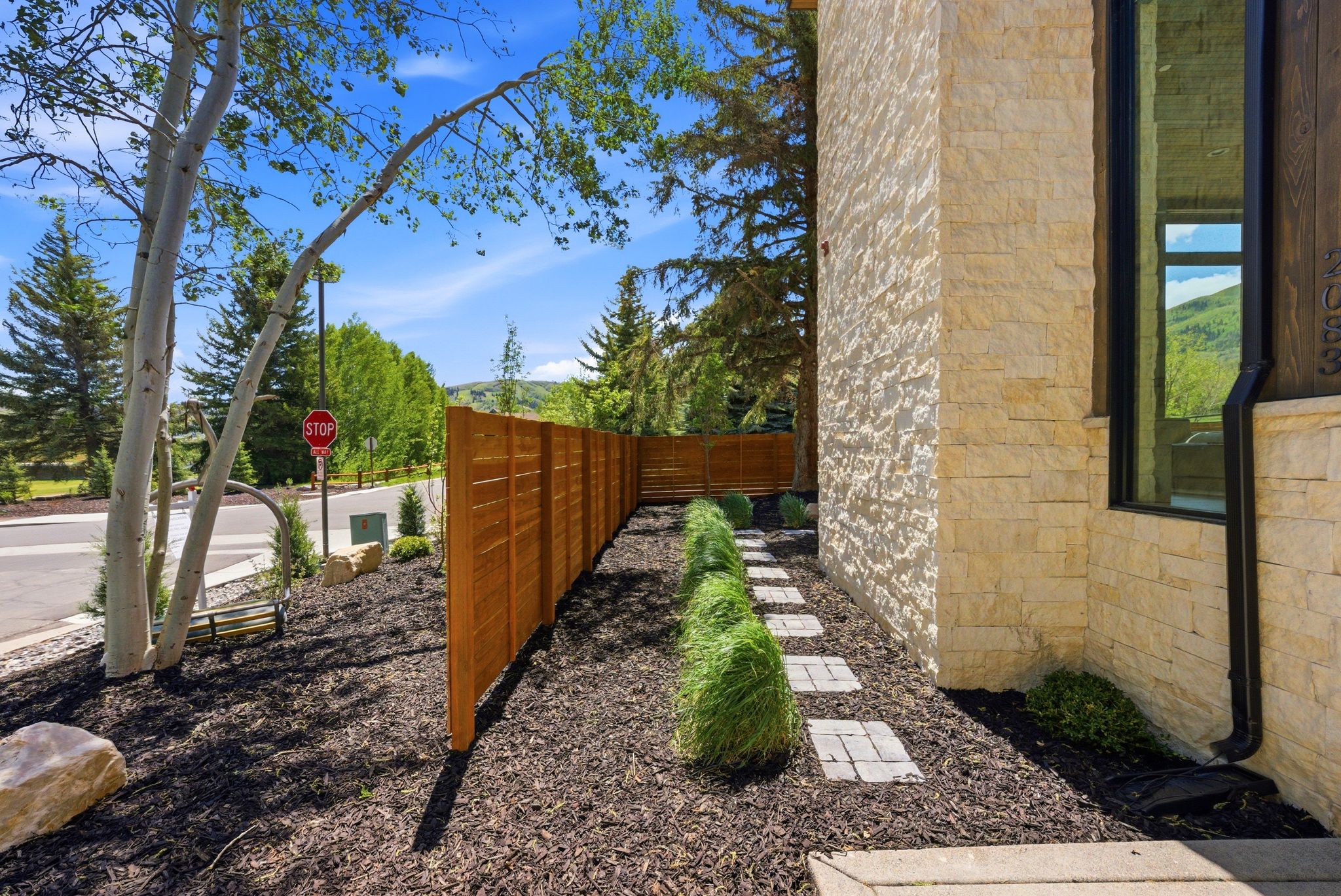 Stone veneer exterior with dark-framed windows, complemented by a wood privacy fence and a landscaped planting bed featuring stepping stones and manicured shrubbery