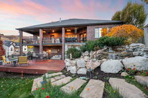 Back of house at dusk featuring a balcony, brick siding, outdoor dining space, roof with shingles, and a deck