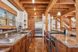 Kitchen with beamed ceiling, brown cabinetry, built in appliances, hanging light fixtures, and glass insert cabinets