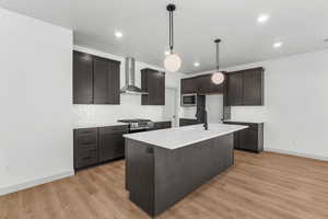 Kitchen with wall chimney exhaust hood, pendant lighting, backsplash, an island with sink, and light wood-type flooring