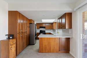 Kitchen featuring wood brown cabinets, a peninsula, stainless refrigerator, black range, and light stone counters