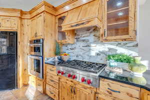 Kitchen featuring decorative backsplash, stainless steel appliances, glass insert cabinets, custom exhaust hood, and dark stone countertops