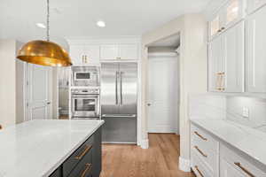 Kitchen with light stone counters, white cabinets, built in appliances, light wood-style flooring, and recessed lighting