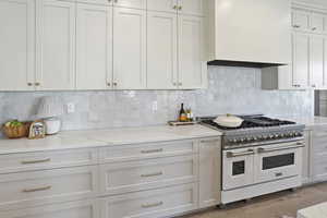Kitchen featuring double oven range, light stone counters, tasteful backsplash, custom range hood, and white cabinets