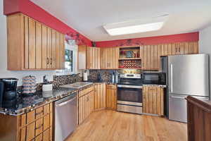 Kitchen with appliances with stainless steel finishes, light wood-type flooring, open shelves, tasteful backsplash, and dark stone counters
