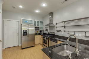 Kitchen featuring appliances with stainless steel finishes, dark stone countertops, wall chimney range hood, glass insert cabinets, and light wood finished floors