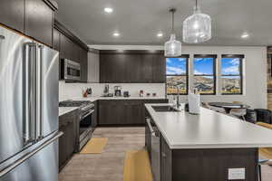 Kitchen with high end appliances, dark brown cabinets, light countertops, light wood finished floors, and a kitchen island with sink