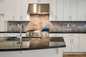 Kitchen with white cabinets, dark stone countertops, under cabinet range hood, decorative backsplash, and stainless steel gas range oven
