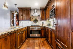 Kitchen featuring hanging light fixtures, light stone countertops, wall chimney range hood, appliances with stainless steel finishes, and backsplash