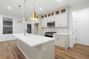 Kitchen featuring decorative backsplash, appliances with stainless steel finishes, a kitchen island with sink, glass insert cabinets, and light stone countertops
