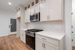 Kitchen featuring appliances with stainless steel finishes, glass insert cabinets, tasteful backsplash, light stone counters, and light wood-style flooring