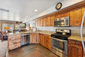 Kitchen featuring stainless steel appliances, a peninsula, brown cabinetry, light stone counters, and open floor plan