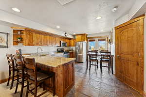 Kitchen featuring brown cabinets, stainless steel appliances, light stone counters, a peninsula, and recessed lighting