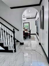 Hallway with ornamental molding, light marble finish flooring, arched walkways, a chandelier, and stairs