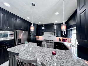 Kitchen featuring dark cabinets, light stone countertops, stainless steel appliances, decorative backsplash, and a kitchen bar