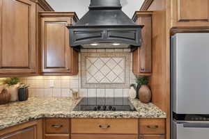 Kitchen featuring backsplash, light stone countertops, custom exhaust hood, freestanding refrigerator, and brown cabinets