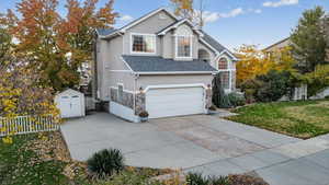 Traditional home featuring a garage, stucco siding, a storage unit, a shingled roof, and driveway