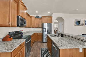Kitchen with stainless steel appliances, brown cabinetry, light stone counters, dark wood-style floors, and arched walkways