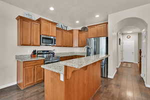 Kitchen with light stone countertops, a kitchen island with sink, brown cabinetry, stainless steel appliances, and arched walkways