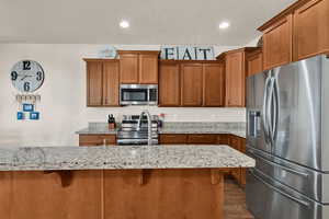 Kitchen featuring appliances with stainless steel finishes, brown cabinets, light stone counters, a kitchen breakfast bar, and recessed lighting