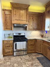 Kitchen with stainless steel gas range oven, brown cabinetry, light stone counters, and range hood