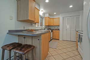 Kitchen with a kitchen bar, light tile patterned flooring, white appliances, light brown cabinets, and a peninsula