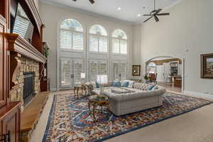 Living area featuring a ceiling fan, carpet, a towering ceiling, ornamental molding, and a stone fireplace