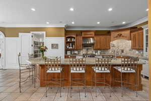 Kitchen with brown cabinetry, a kitchen bar, light stone counters, crown molding, and light tile patterned floors