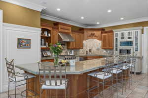 Kitchen featuring brown cabinetry, tasteful backsplash, a breakfast bar area, wall chimney range hood, and light tile patterned floors