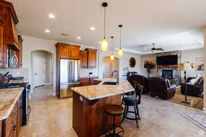 Kitchen featuring arched walkways, appliances with stainless steel finishes, hanging light fixtures, a kitchen island with sink, and brown cabinetry