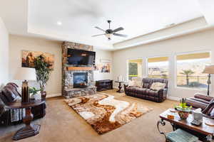 Living room featuring a tray ceiling, light carpet, ceiling fan, and a stone fireplace