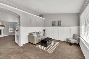 Living area with a decorative wall, wainscoting, carpet, ceiling fan, and a fireplace with raised hearth